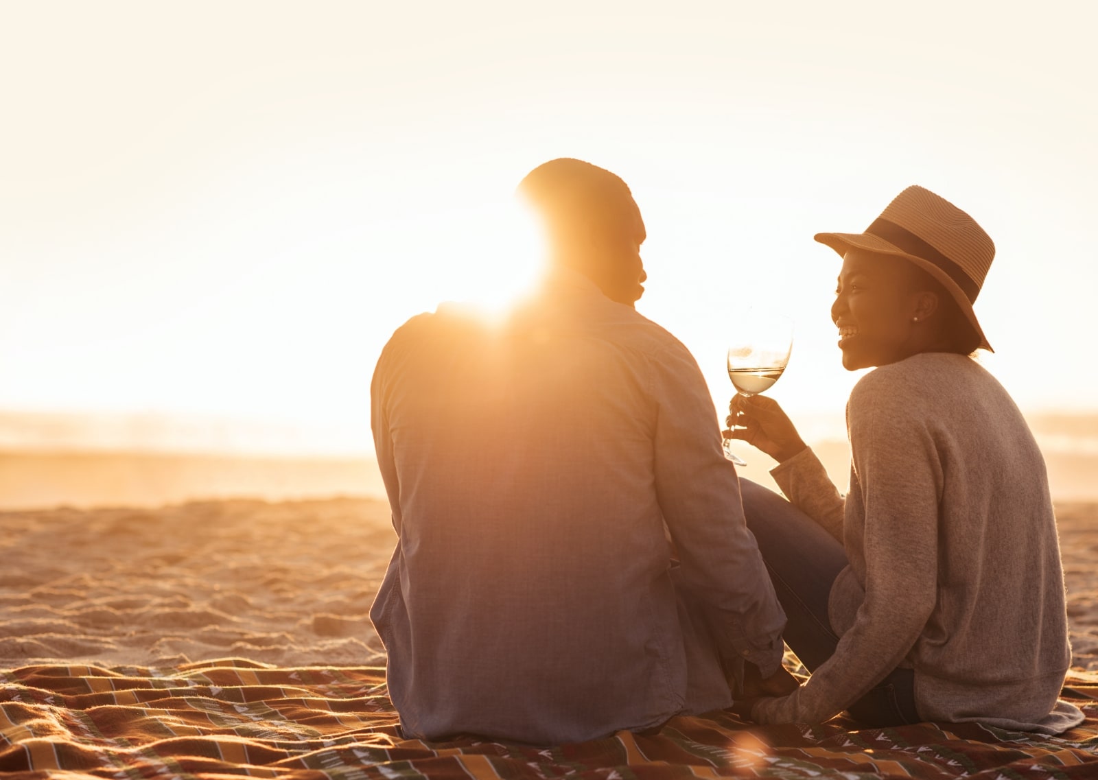 a man and woman sitting on a beach at sunset drinking wine